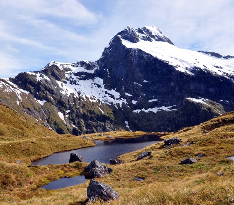 Snow-capped mountain behind a small alpine lake with grassy and rocky terrain.