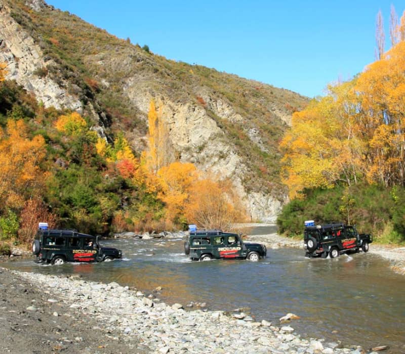 Three offroad vehicles driving through a shallow river with autumn trees and rocky hills around.