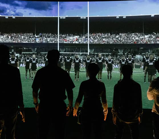 Silhouettes watching an All Blacks rugby match on a large screen.