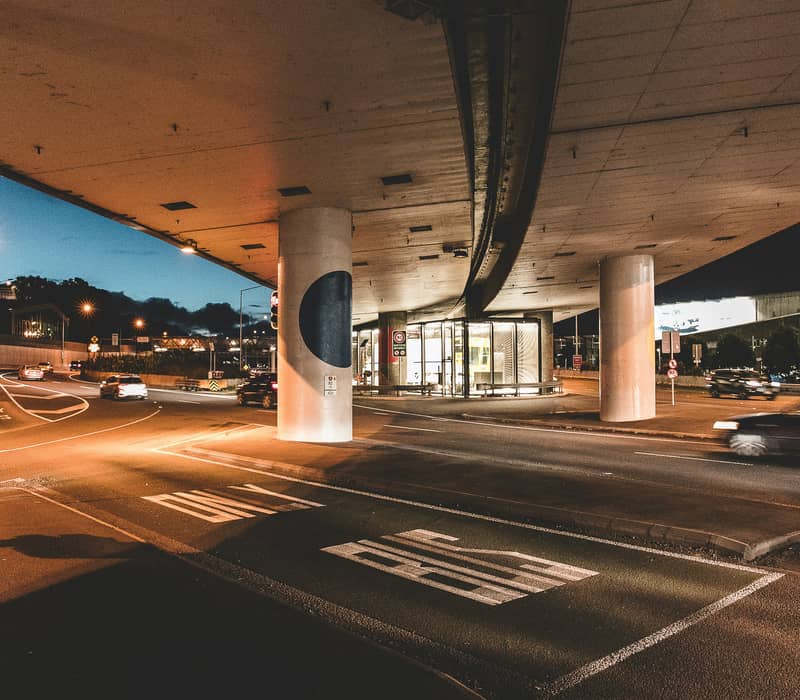 View from under a concrete highway overpass at night showing a city intersection with streetlights, road signs, and motion-blurred cars.