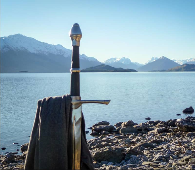 Sword with scarf on rocky shore by lake with snow-capped mountains.