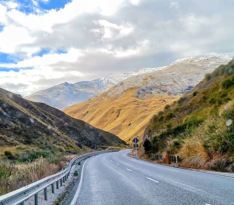 Winding mountain road through grassy hills and snow-capped peaks under a cloudy blue sky.