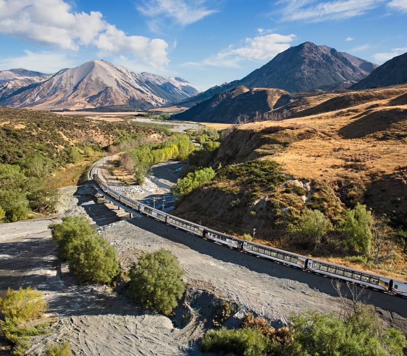 TranzAlpine train crossing a river bridge in a mountainous valley under blue sky and clouds
