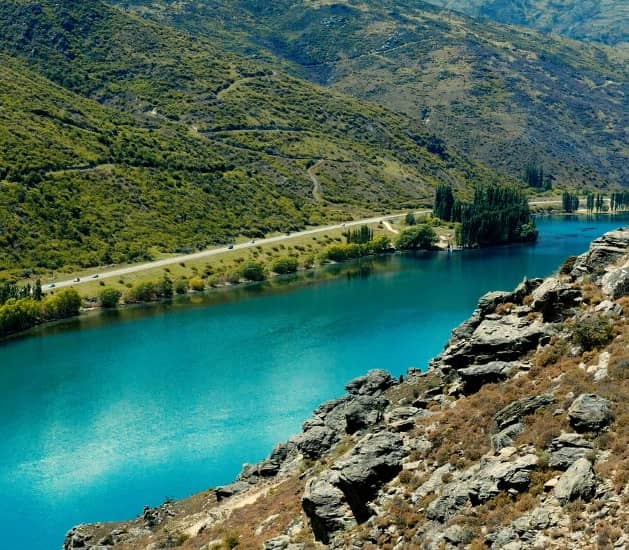 Blue river flowing beside a winding road with green hills and rocky cliffs.