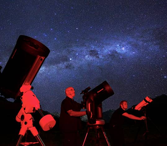 Three people using telescopes to stargaze under a starry night sky with the Milky Way visible.