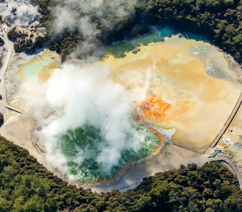 Aerial shot of a colorful geothermal hot spring emitting steam with forest and boardwalks nearby