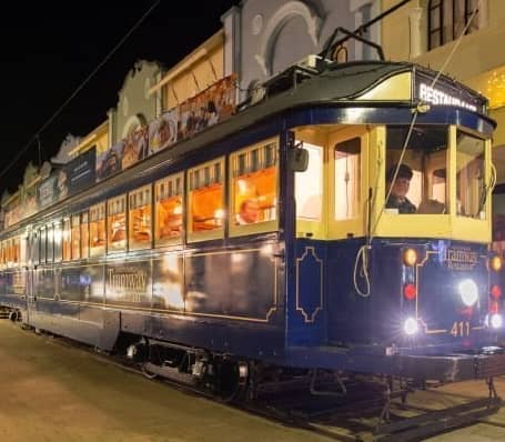 Historic blue tram with glowing interior lights traveling on a city street at night.