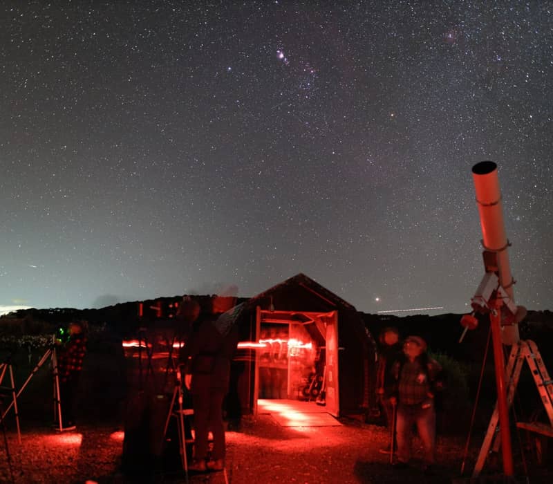 People using telescopes to view stars under the Milky Way with a red-lit observatory tent at night.