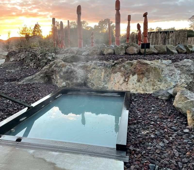 Steaming hot spring pool with wooden statues and rocky landscape at sunset.