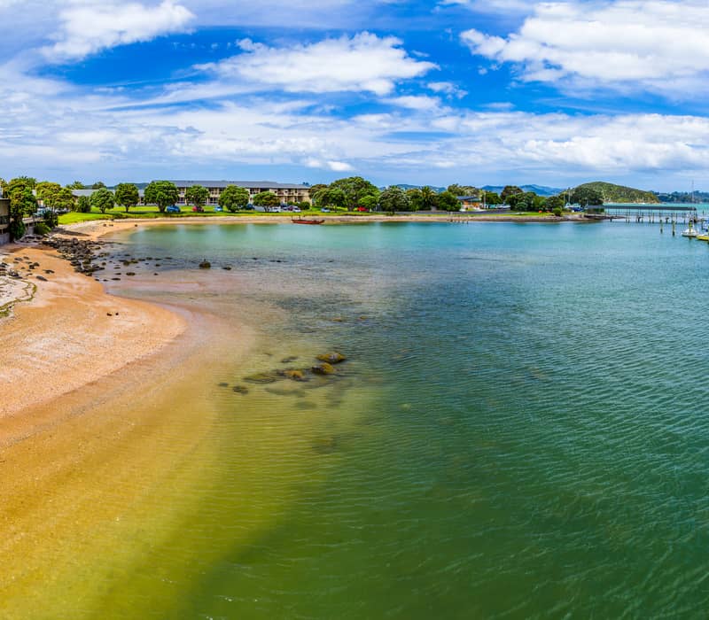 A panoramic view of Te Ti Bay in Paihia, showing a golden sand beach, clear green-blue water with moored boats, and beachfront buildings under a bright blue sky.