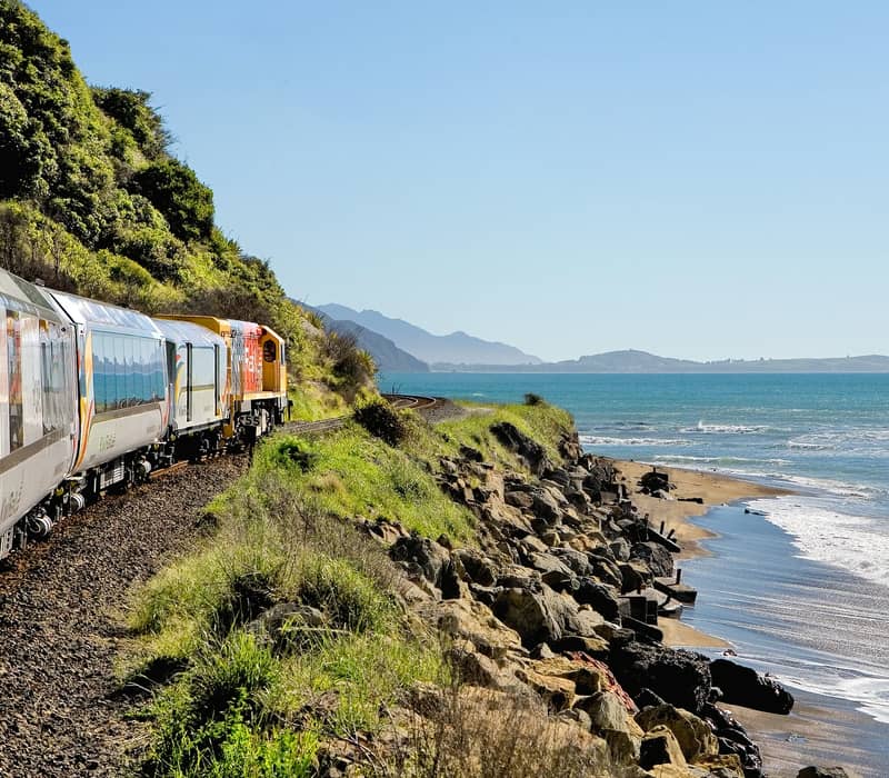 Train traveling along a coastal railway with the ocean and beach nearby under a clear sky.
