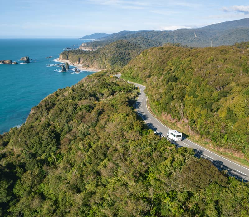 An aerial view of a white campervan driving along a winding coastal road bordered by dense green rainforest and the turquoise Tasman Sea.