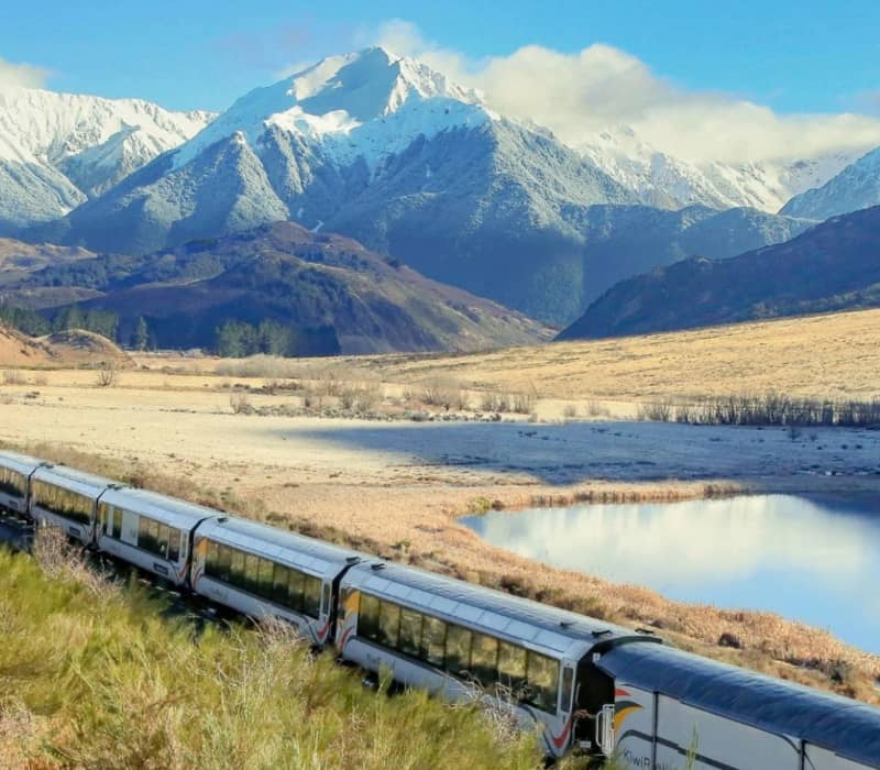 Train traveling through a mountain valley with snow-covered peaks and a calm lake.