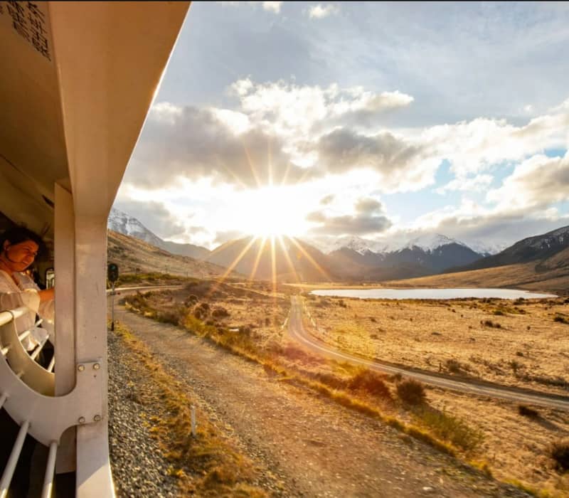 People on a train looking at a sunset over mountains and a lake in a wide valley.