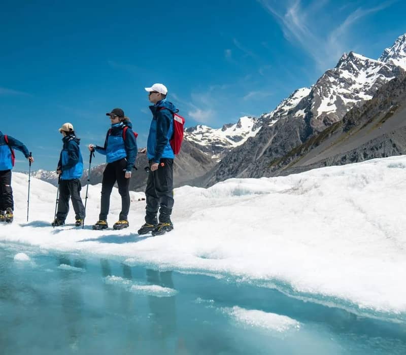 Group of hikers trekking on a glacier with snow and mountains in the background.