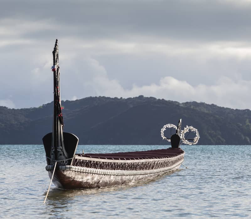 Traditional carved waka boat on calm water with hills and cloudy sky in the background Credit: Waitangi Treaty Grounds
