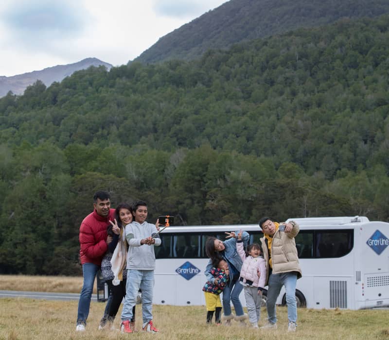 Group of tourists taking selfies in a grassy valley with a Gray Line bus and forested mountains behind them