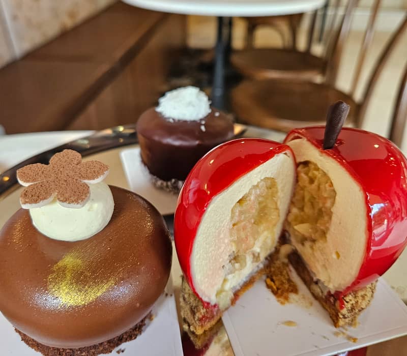 Three artistic cakes on display with one red apple-shaped cake cut open on a reflective table inside a cafe.