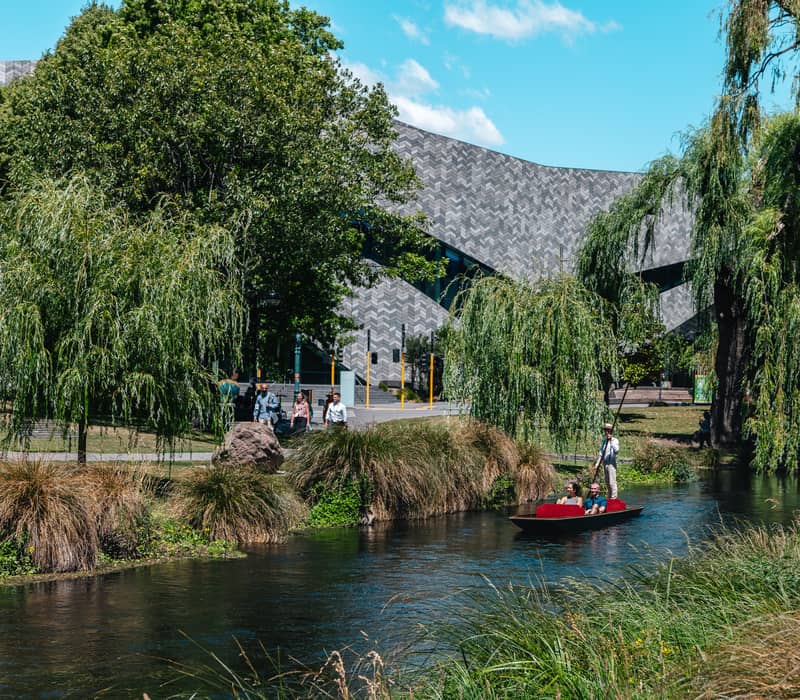 People in a punt boat on a river with trees, grass, and a modern building in the background under blue sky.