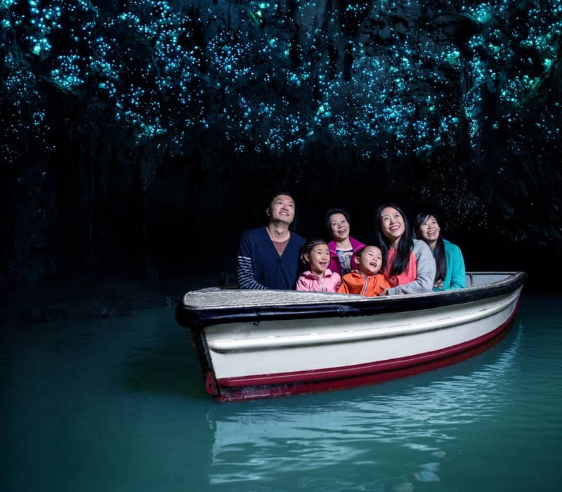 Family on boat ride inside a dark cave illuminated by glowing blue glowworms above.