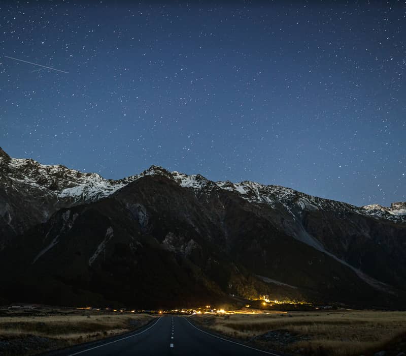 An empty road leads toward the glowing lights of Mount Cook Village at the base of dark, snow-dusted mountains under a vast, star-filled night sky. Credit: New Zealand Fine Touring