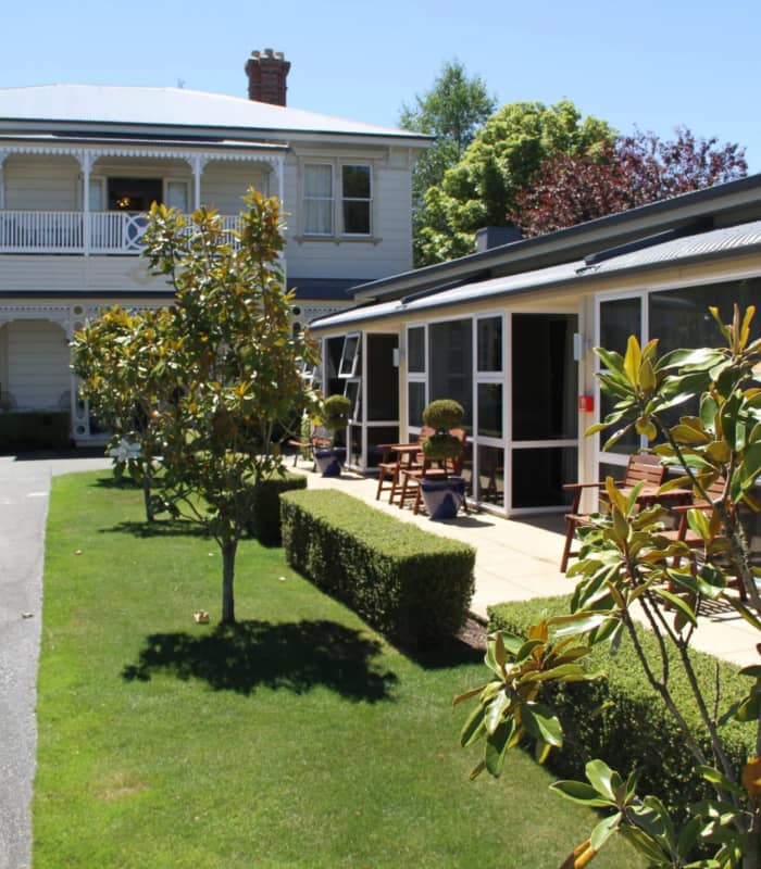 Two-story manor building with garden, flowering plants, and benches outside on a sunny day.