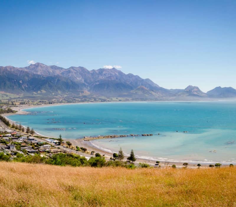 Coastal bay with clear blue water, town by the shore, mountains in background under a blue sky.