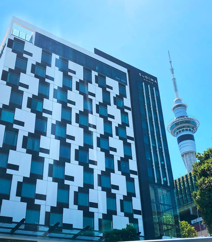 Sudima Auckland hotel building with a modern checkered design and the Sky Tower nearby under a blue sky.