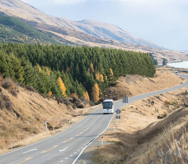 Coach driving on winding road next to Lake Pukaki with hills and trees showing autumn colors