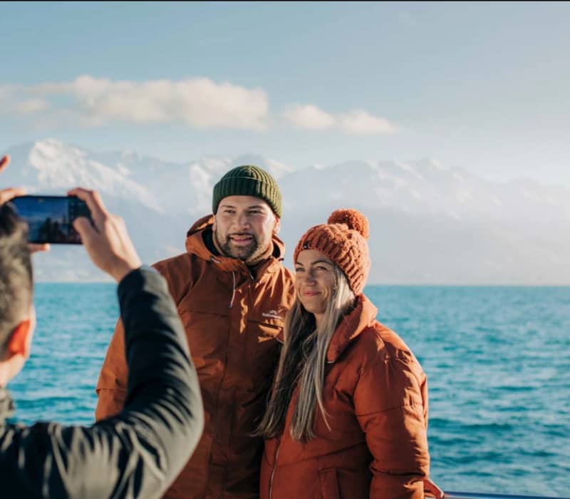 Couple in warm jackets and beanies posing by a lake with snowy mountains behind.