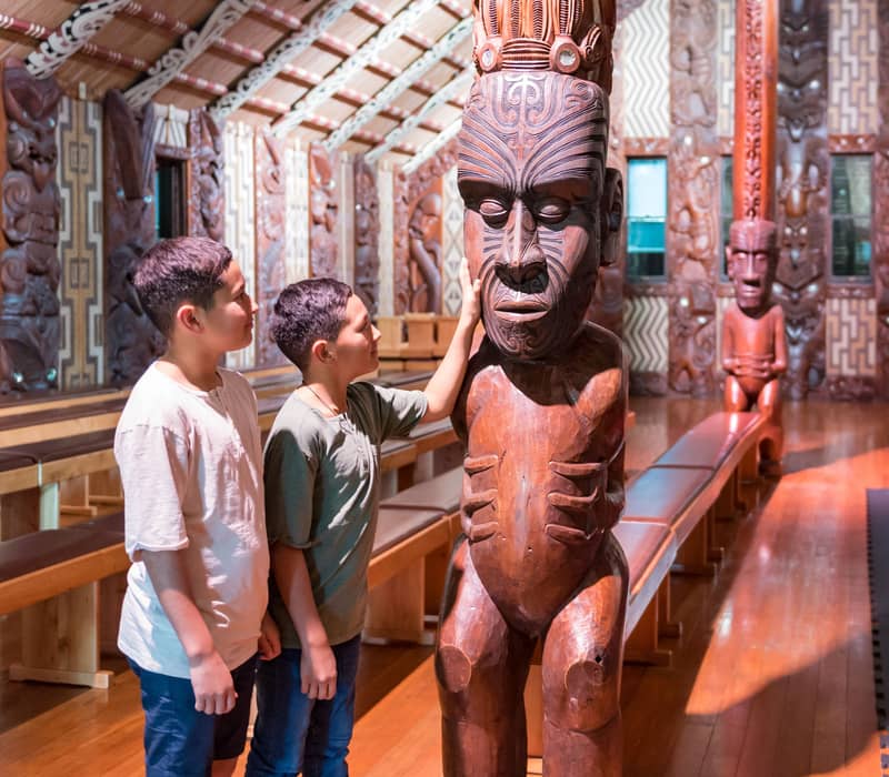 Two boys looking at and touching a large carved wooden Maori statue inside a decorated hall.