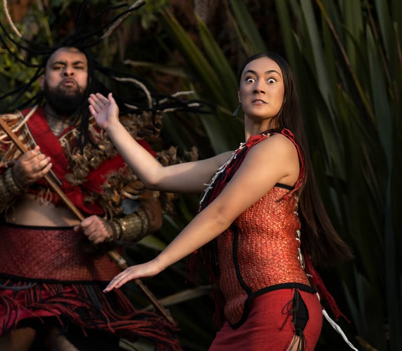 Two Maori performers performing a traditional dance with expressive faces and traditional attire in a natural setting.