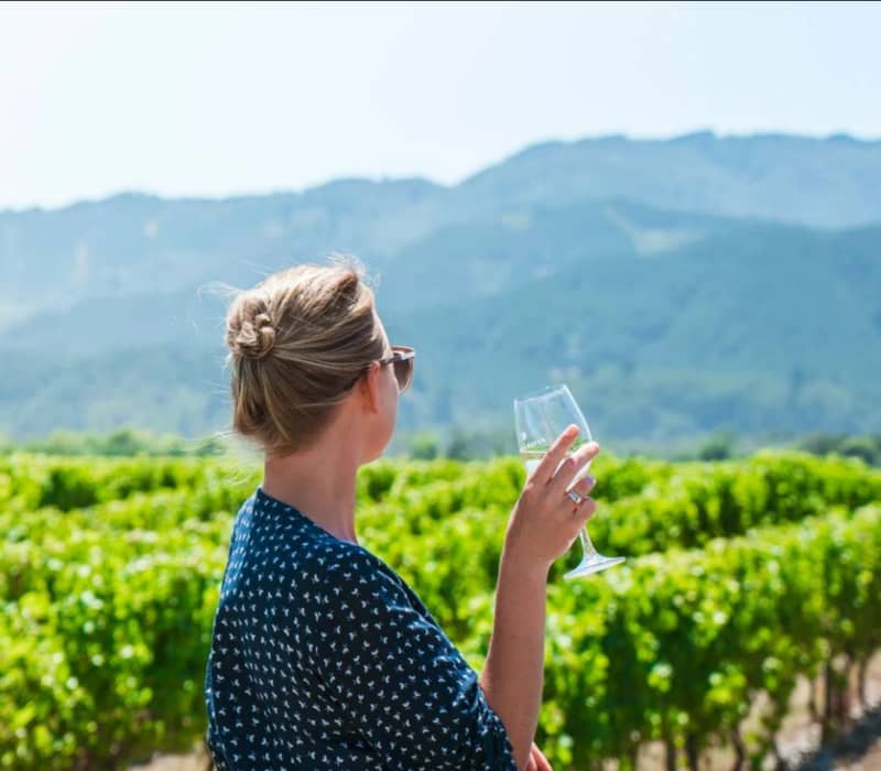 Woman holding a glass of white wine standing in a green vineyard with mountains in the background.