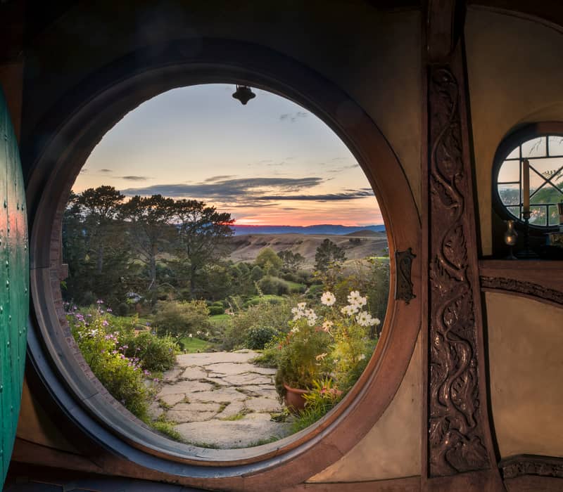 View through a round hobbit door opening to a garden and hills at sunset