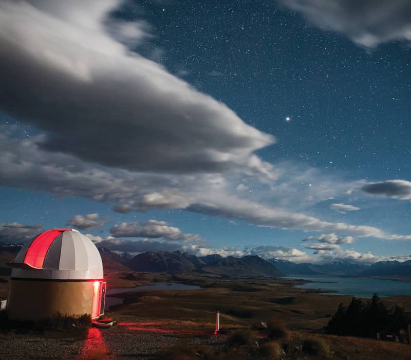 Observatory dome with red light under a starry sky and mountains by a lake at night.