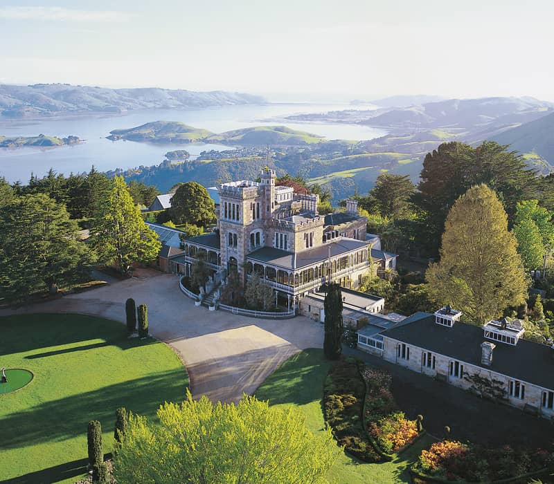 Historic stone castle with gardens, trees, hills, and a lake in the distance under a clear sky.