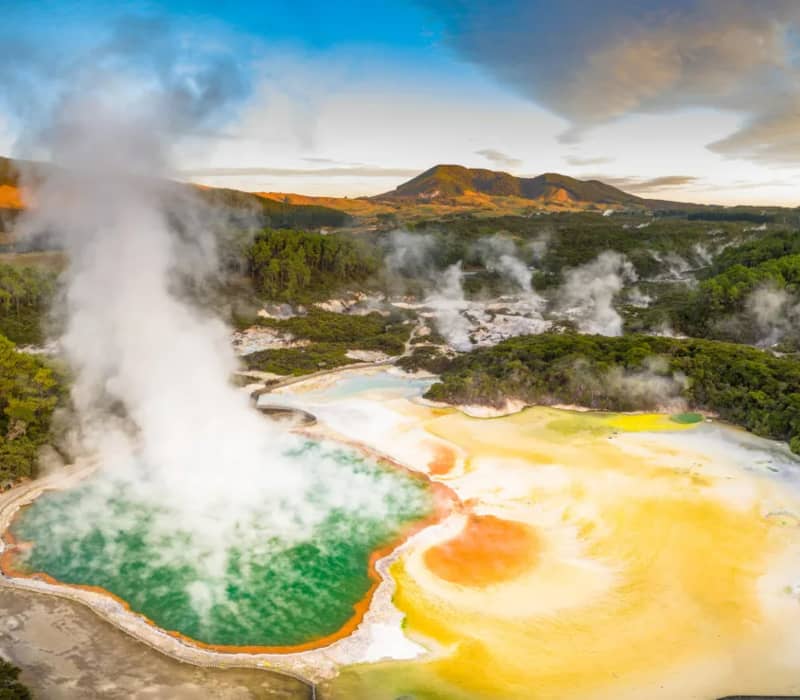 An aerial-style view of the vibrant Champagne Pool at Wai-O-Tapu Thermal Wonderland, with its deep green water, bright orange mineral rim, and a massive plume of white steam rising into the sky.