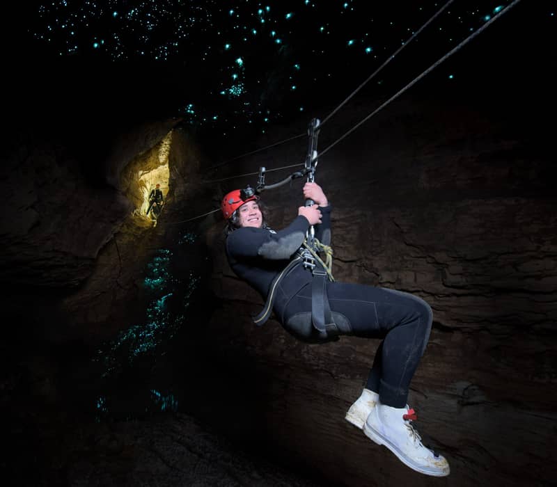 A person wearing a red safety helmet and a wetsuit ziplining through a dark cave, with thousands of tiny blue-green bioluminescent glowworms illuminating the ceiling above them like a starry night.