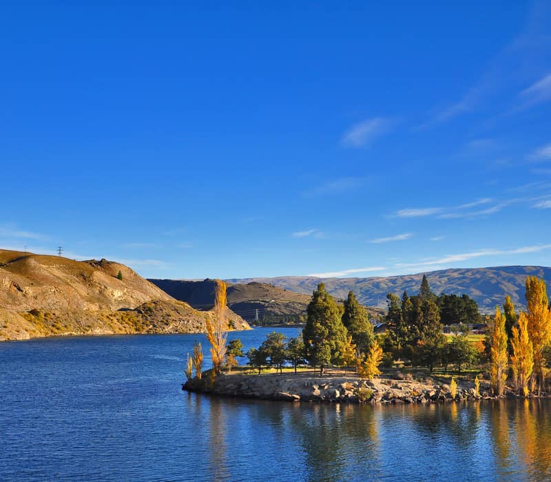 Lake with autumn trees and hills under clear blue sky reflecting on the water surface.