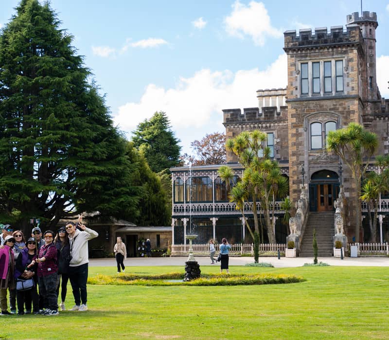 Tourists taking a group selfie near Larnach Castle with garden and trees.