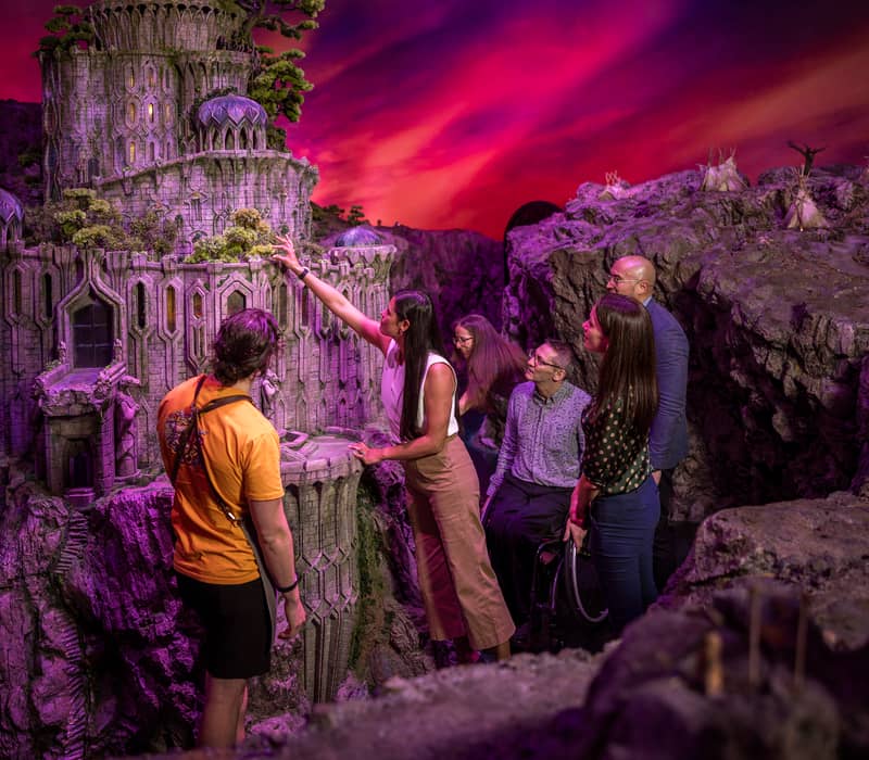 Tour group examines a large detailed castle model under purple lighting indoors
