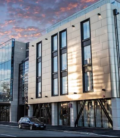 Exterior of a modern hotel building with glass windows reflecting sunset sky and a car parked outside.