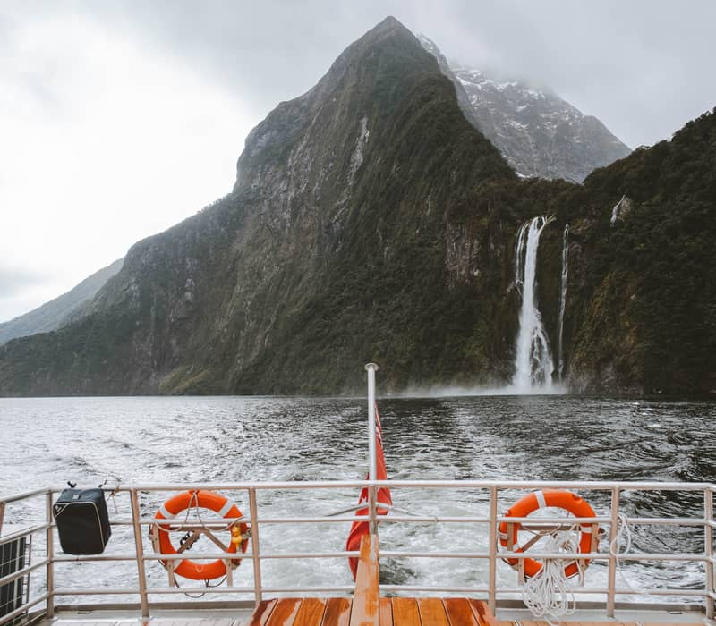 View from boat deck towards waterfalls cascading down a mountain beside a lake under a cloudy sky.