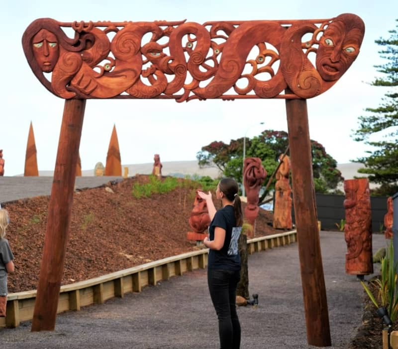 A group of visitors, including children, listening to a Māori guide in front of a monumental, intricately carved wooden gateway at Manea Footprints of Kupe. Credit: Manea Footprints of Kupe