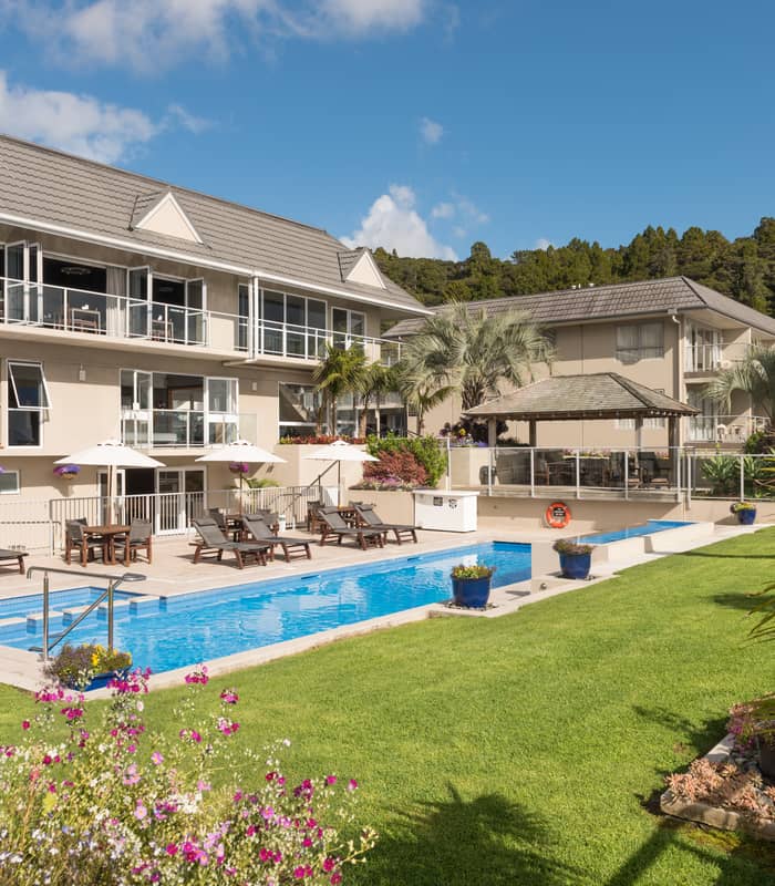 Outdoor pool with lounge chairs and umbrellas in front of a hotel building with balconies under a clear blue sky.