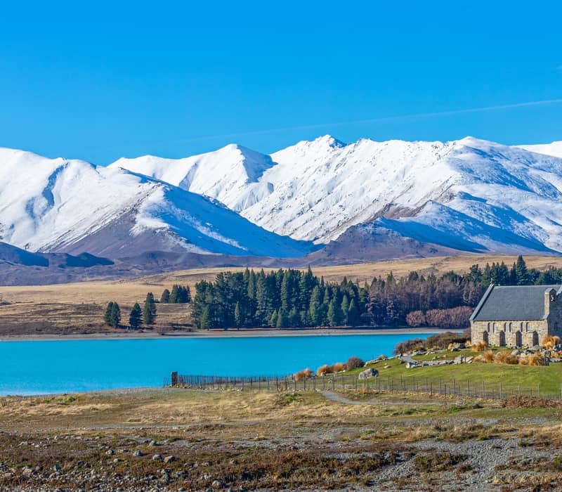 Stone church by bright blue lake with snow-capped mountains in background under clear sky.