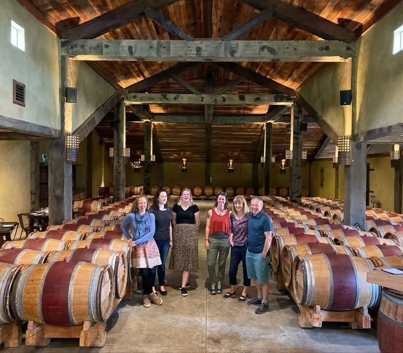 Group of six people standing among rows of wooden wine barrels in a winery tasting room.