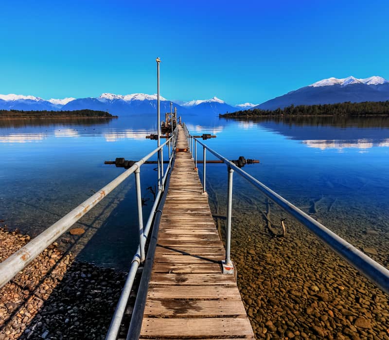 Wooden jetty stretching into a lake with clear water and snowcapped mountains under a bright blue sky.