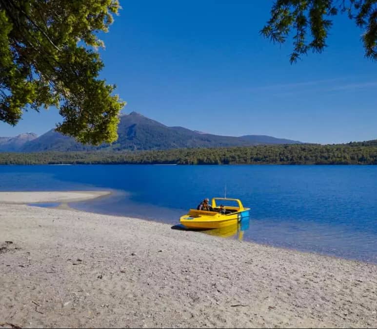 Yellow boat on a lake with sandy beach, forest, and mountains under blue sky.