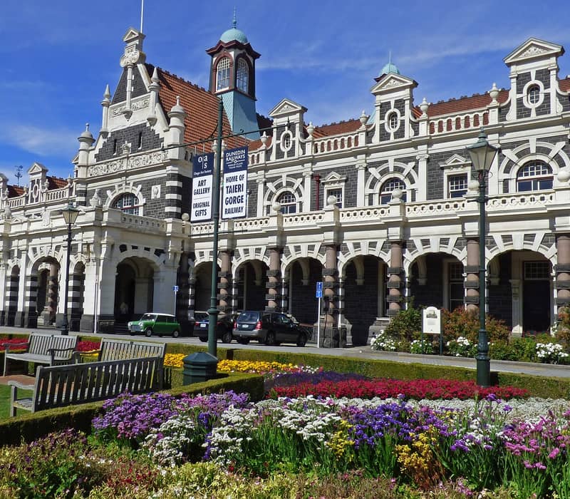 Historic Dunedin Railway Station with flower gardens and benches under a clear blue sky.